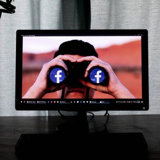man on computer screen looks through binoculars with facebook logos on lenses